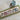 Cross-stitched bookmark with a village scene on a green fabric strip, with a small house model beside it.