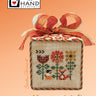 Cross-stitched sampler with floral design and ribbons on a peach background, featuring Heart in Hand Needleworks branding.