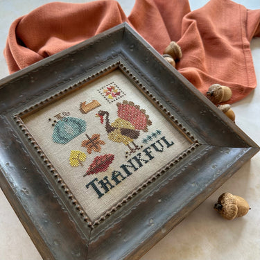Cross-stitched piece with a turkey and 'Thankful' text, framed in a rustic wooden box, on a neutral background.