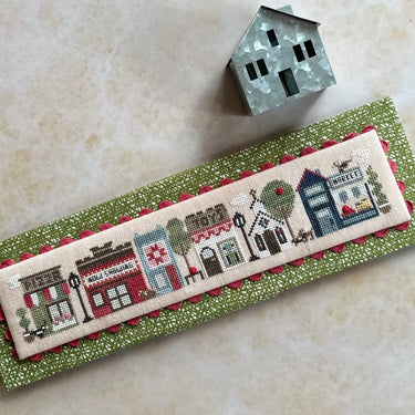 Cross-stitched bookmark with a village scene on a green fabric strip, with a small house model beside it.