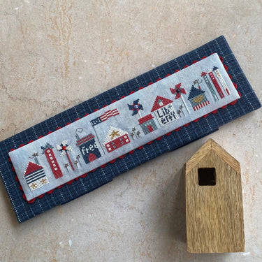 Cross-stitched panel with small buildings and text on a textured surface with a wooden birdhouse.
