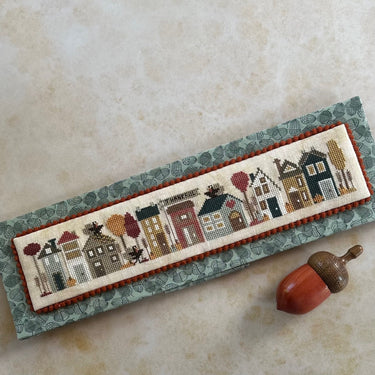 Cross-stitched panel with village scene on a textured surface with an acorn.
