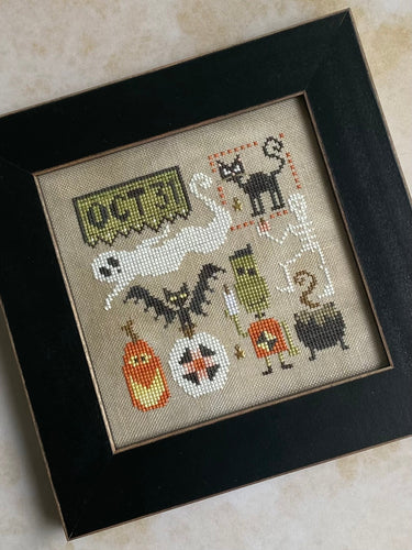 Framed cross-stitched artwork with Halloween-themed designs on a beige background.