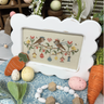 Cross-stitched picture of birds and eggs in a white frame on a table with Easter decorations.