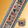 Cross-stitched pattern of a tiny town with a small stool featuring the same design, on a yellow background.